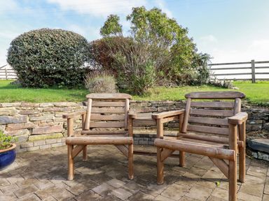 A patio with two wooden chairs connected by a small table and a stone wall with bushes behind at Great Meadow Barn in Crantock