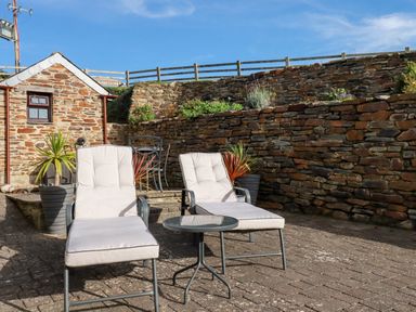 An outdoor patio area with two cushioned lounge chairs and a small table near a stone wall and a small stone building at Great Meadow Barn in Crantock