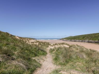 A sandy path through grassy dunes leading to a wide sandy beach with hills and ocean in the background at Great Meadow Barn in Crantock