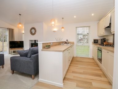 A kitchen with light wood floors and cream cabinets next to a living area with grey sofas and a TV at Hollytree Lodge Beckside 1 Limefitt Park near Windermere