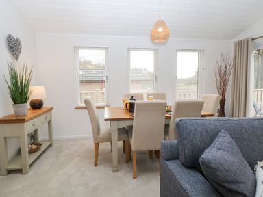 A dining area with a wooden table and upholstered chairs next to a side table with a lamp and plant at Hollytree Lodge Beckside 1 Limefitt Park near Windermere