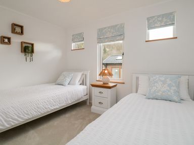 A bedroom with two single beds with white bedding a wooden nightstand with a lamp and three small windows with blue floral blinds at Hollytree Lodge Beckside 1 Limefitt Park near Windermere