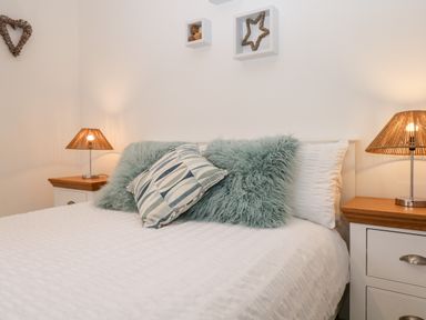 A bedroom with a bed with white bedding two light blue fluffy pillows and one patterned pillow two bedside tables with lamps a heart shaped wall hanging and square wall shelves at Hollytree Lodge Beckside 1 Limefitt Park near Windermere