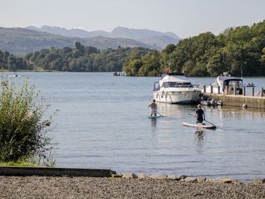 Two people paddleboarding on a lake near boats and a dock with hills in the background at Hollytree Lodge Beckside 1 Limefitt Park near Windermere