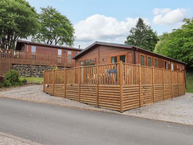 A wooden lodge with a fenced deck and gravel driveway surrounded by trees at Hollytree Lodge Beckside 1 Limefitt Park near Windermere