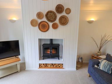 A living room with a black stove in a white fireplace surrounded by woven baskets a TV on a wooden stand and a blue sofa with a cushion at Hollytree Lodge Beckside 1 Limefitt Park near Windermere