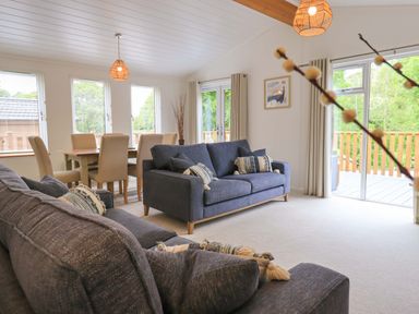 A living room with dark sofas a dining table with beige chairs and large windows at Hollytree Lodge Beckside 1 Limefitt Park near Windermere