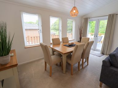 A dining room with six beige chairs around a wooden table near windows and glass doors at Hollytree Lodge Beckside 1 Limefitt Park near Windermere