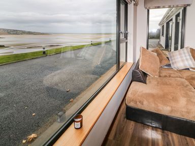 A corner of a room with a brown sectional sofa and cushions next to large windows overlooking a road and a beach at Upper Killult in Killult near Gortahork County Donegal