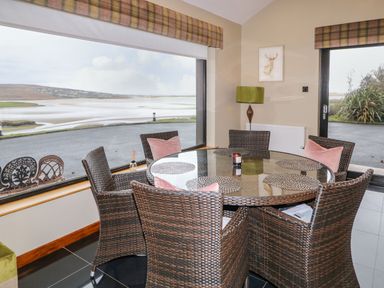 A dining area with a round glass table and wicker chairs with pink cushions near large windows overlooking a water view at Upper Killult in Killult near Gortahork County Donegal