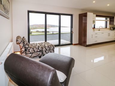 A room with patterned armchairs, a dark leather chair, a kitchen area, and large sliding glass doors showing a water view at Upper Killult in Killult near Gortahork County Donegal