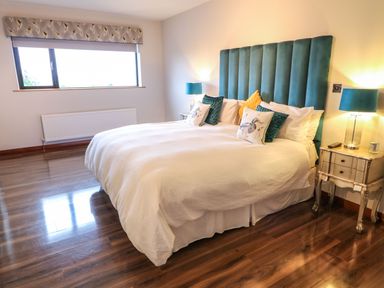 A bedroom with a large bed with white bedding and teal headboard two bedside tables with lamps and a window with a valance at Upper Killult in Killult near Gortahork County Donegal