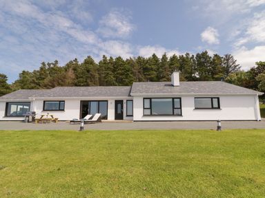 A single story white house with large windows and outdoor seating on a lawn surrounded by trees at Upper Killult in Killult near Gortahork County Donegal