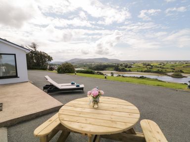 A patio area with a round wooden picnic table with flowers and a white lounge chair with blue towels overlooking a river and green fields at Upper Killult in Killult near Gortahork County Donegal
