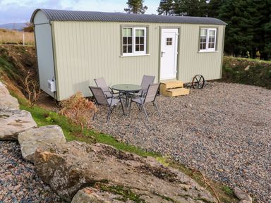 An outdoor area with a small building, a round glass table with four chairs, gravel ground and large rocks nearby at Shepherds Retreat Glenlark in Gortin near Greencastle