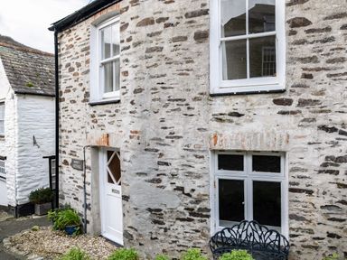 Exterior view of a stone cottage with white framed windows a white door and a metal bench at Honey Cottage in Port Isaac