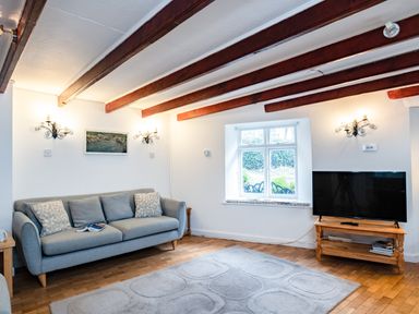 A living room with a grey sofa a wooden TV stand with a television a window and wooden ceiling beams at Honey Cottage in Port Isaac