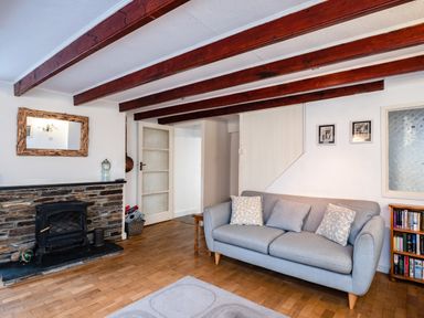 A living room with a gray sofa a stone fireplace and wooden ceiling beams at Honey Cottage in Port Isaac