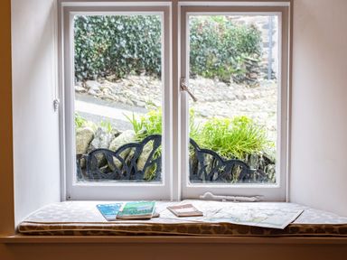 A window with a cushioned seat holding maps and booklets overlooking a garden with stones and greenery at Honey Cottage in Port Isaac