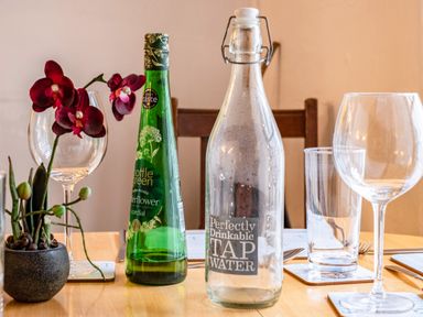 A dining table with a bottle of elderflower cordial a bottle of tap water drinking glasses and a small pot with red flowers at Honey Cottage in Port Isaac