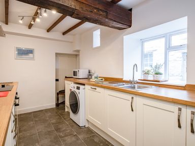 A kitchen with white cabinets a washing machine a microwave and a sink under a window at Honey Cottage in Port Isaac
