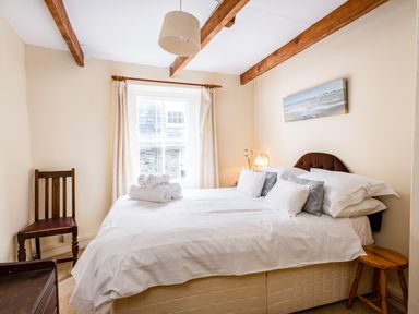 A bedroom with a bed with white linens pillows and towels near a window with curtains and wooden beams on the ceiling at Honey Cottage in Port Isaac