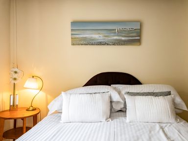A bedroom with a bed with white pillows and striped white bedding a small wooden side table with a lamp and flowers on it and a painting above the bed at Honey Cottage in Port Isaac