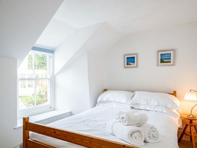 A bedroom with a wooden bed with white linens and towels next to a window and a small lamp on a side table at Honey Cottage in Port Isaac