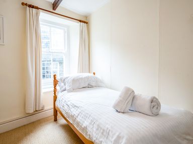 A bedroom with a single bed white striped bedding two pillows and two rolled towels near a window with light curtains at Honey Cottage in Port Isaac