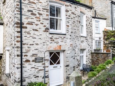 The exterior of a stone cottage with white window frames and a white door with a sign reading honey cottage in port isaac