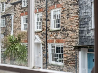 View of a stone house with white framed windows and a white door seen through a window at Honey Cottage in Port Isaac