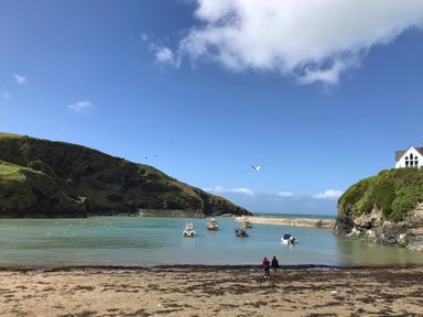 A beach with two people standing on sand near the water where several boats are anchored and hills and a house are in the background at Honey Cottage in Port Isaac