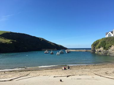A beach with two people sitting on the sand near boats in the water and hills on each side at Honey Cottage in Port Isaac