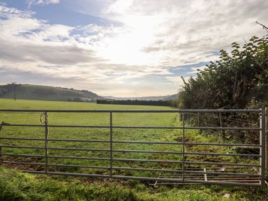 A metal gate opening to a green field with hills and cloudy sky at 3 Caddaford Barns in Buckfastleigh