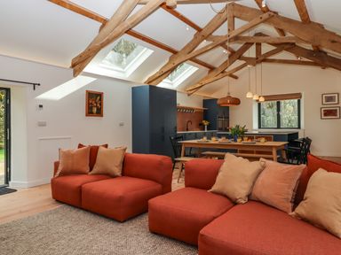 A living room with two red sofas and beige pillows with an open kitchen and dining area under wooden ceiling beams at 3 Caddaford Barns in Buckfastleigh