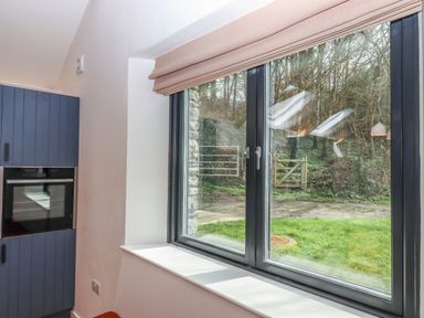 A kitchen area with blue cabinets and an oven near a window showing a garden with a wooden gate at 3 Caddaford Barns in Buckfastleigh