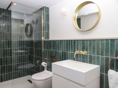 A bathroom with green tiles a glass shower enclosure a white toilet a white sink with gold fixtures and a round gold framed mirror at 3 Caddaford Barns in Buckfastleigh