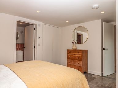 A bedroom with a bed covered by a beige blanket a wooden chest of drawers with a round mirror above and two open doors at 3 Caddaford Barns in Buckfastleigh