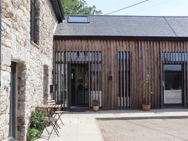 An outdoor patio area with a wooden table and chairs next to a stone building and a wooden slatted facade at 3 Caddaford Barns in Buckfastleigh