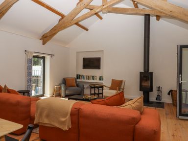 A living room with exposed wooden beams an orange sofa armchairs a wood stove and a bookshelf at 3 Caddaford Barns in Buckfastleigh