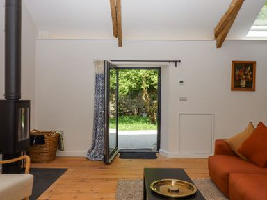 A living room with a wood stove next to a basket open doors showing a garden an orange sofa with cushions and a coffee table with a tray at 3 Caddaford Barns Buckfastleigh