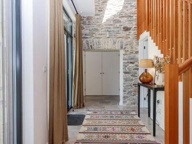 A hallway with stone walls a patterned rug curtains a wooden staircase and a table with a lamp and dried flowers at 3 Caddaford Barns in Buckfastleigh