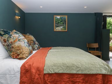 A bedroom with a bed covered with green and orange bedding and patterned pillows a chair and a painting on the wall at 3 Caddaford Barns in Buckfastleigh