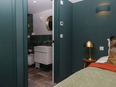 A bedroom corner with a bed a side table and a lamp with an open doorway showing a bathroom with a sink mirror and toilet at 3 Caddaford Barns in Buckfastleigh