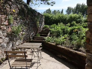 An outdoor seating area with wooden chairs and a table next to stone walls and garden steps at 3 Caddaford Barns in Buckfastleigh