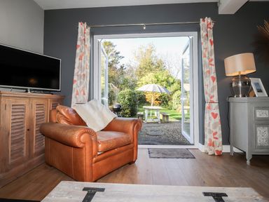 A living room with a leather armchair a wooden cabinet with a television curtains and an open door leading to a garden with a table and umbrella at The Croft in Newport Pembrokeshire