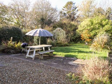An outdoor garden area with a wooden picnic table, umbrella, barbecue grill, and surrounding greenery at The Croft in Newport Pembrokeshire
