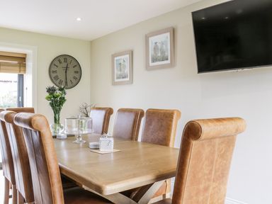 A dining room with a wooden table, brown leather chairs, a wall clock, two framed pictures, a mounted television, and a vase with flowers at Partridge Place in Burton Pidsea