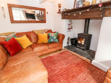 A living room with a leather sofa, colorful cushions, a wood-burning stove, and a large wall mirror at Honeysuckle Cottage in St Agnes