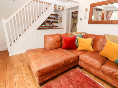 A living room with a brown leather sectional sofa with colorful cushions under a white staircase with shelves at Honeysuckle Cottage in St Agnes